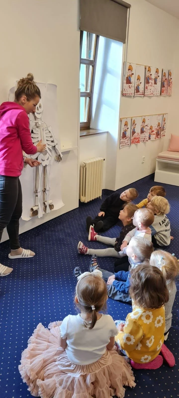 A woman in a pink hoodie is teaching a lesson about the human skeleton to a group of young children seated on a blue carpeted floor in a classroom. The children are looking at a life-sized skeleton model on the wall, and there are colorful drawings on the wall behind them.