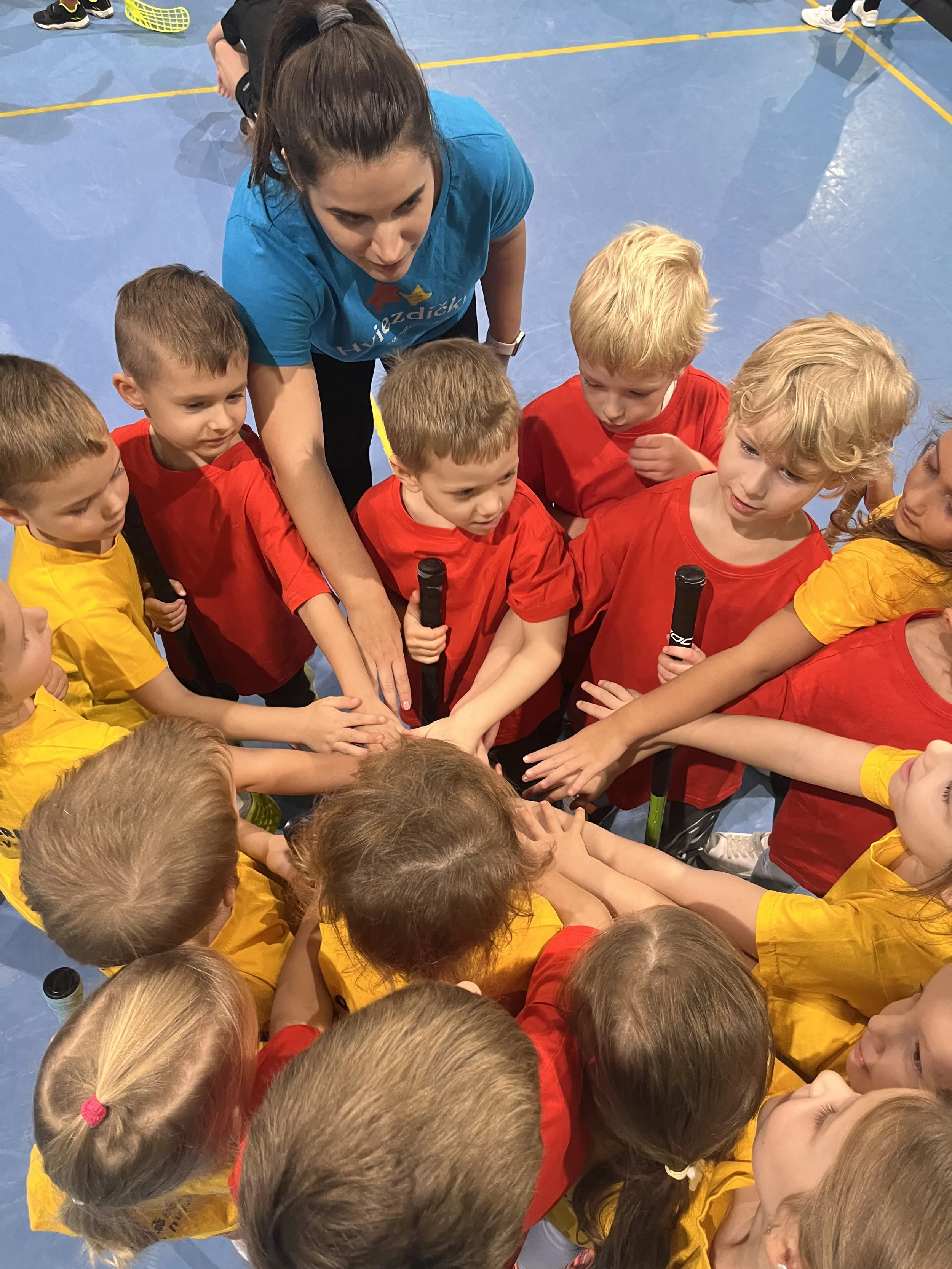 Children in red and yellow team uniforms gather around a female coach in a blue shirt on a sports court. They all place one hand on top of each other's in a team huddle, some holding microphones.