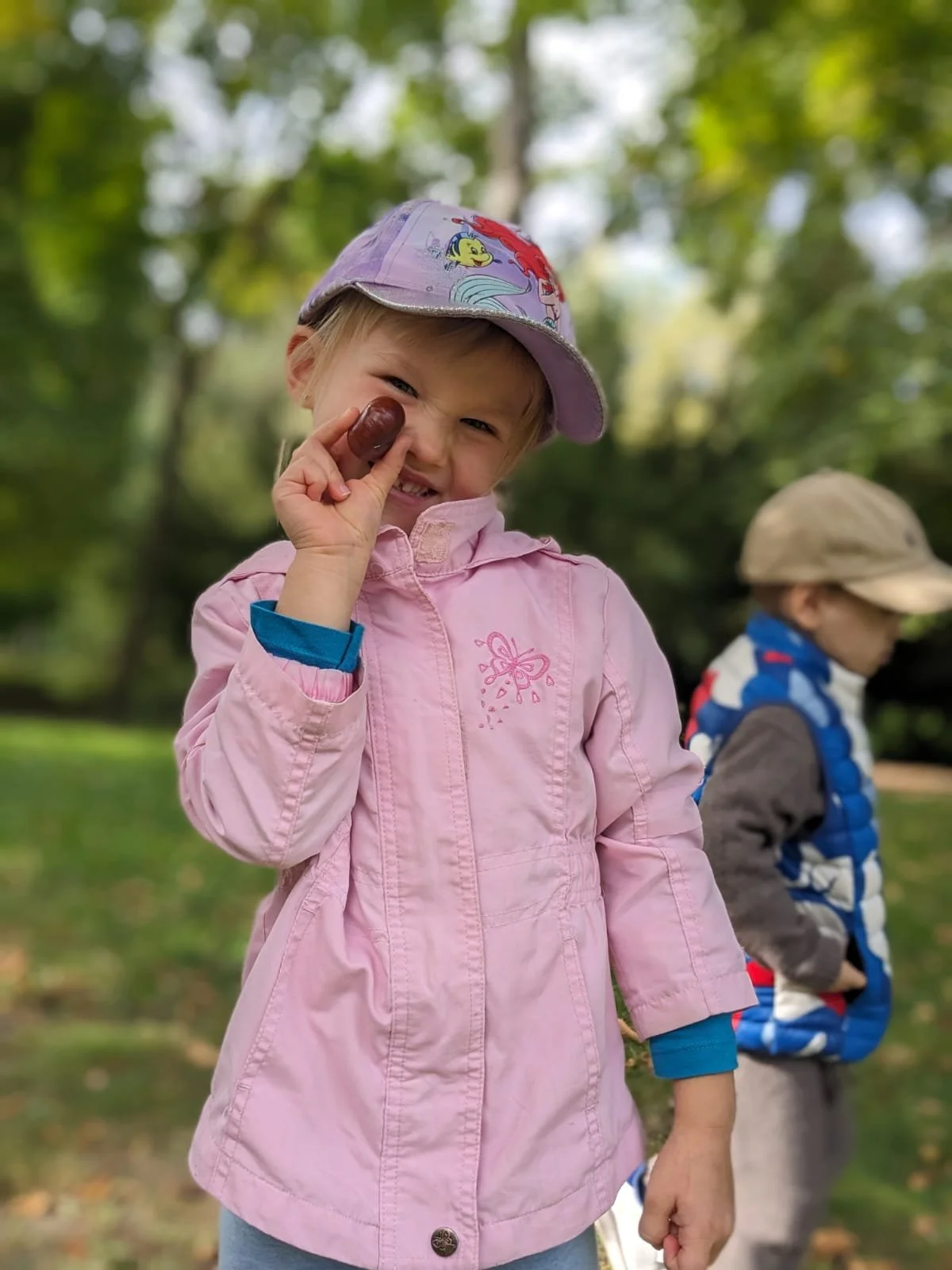 A young girl in a pink jacket and purple hat holding a small brown object, smiling cheerfully, outdoors in a wooded area with a boy wearing a beige cap and blue vest in the background.