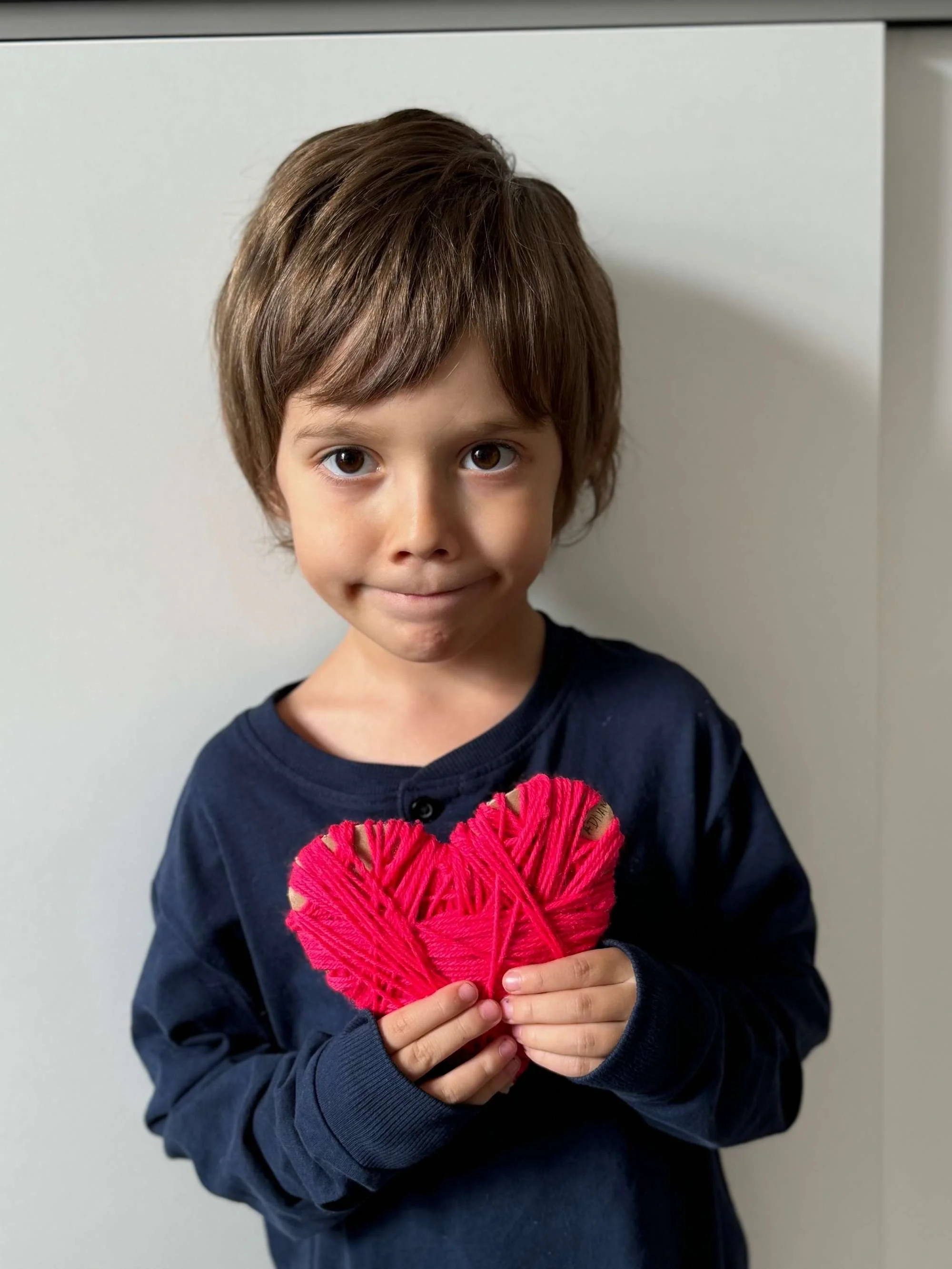 A young boy with brown hair and brown eyes, wearing a navy blue shirt, holding a handmade pink heart-shaped craft made from yarn, standing against a plain background.