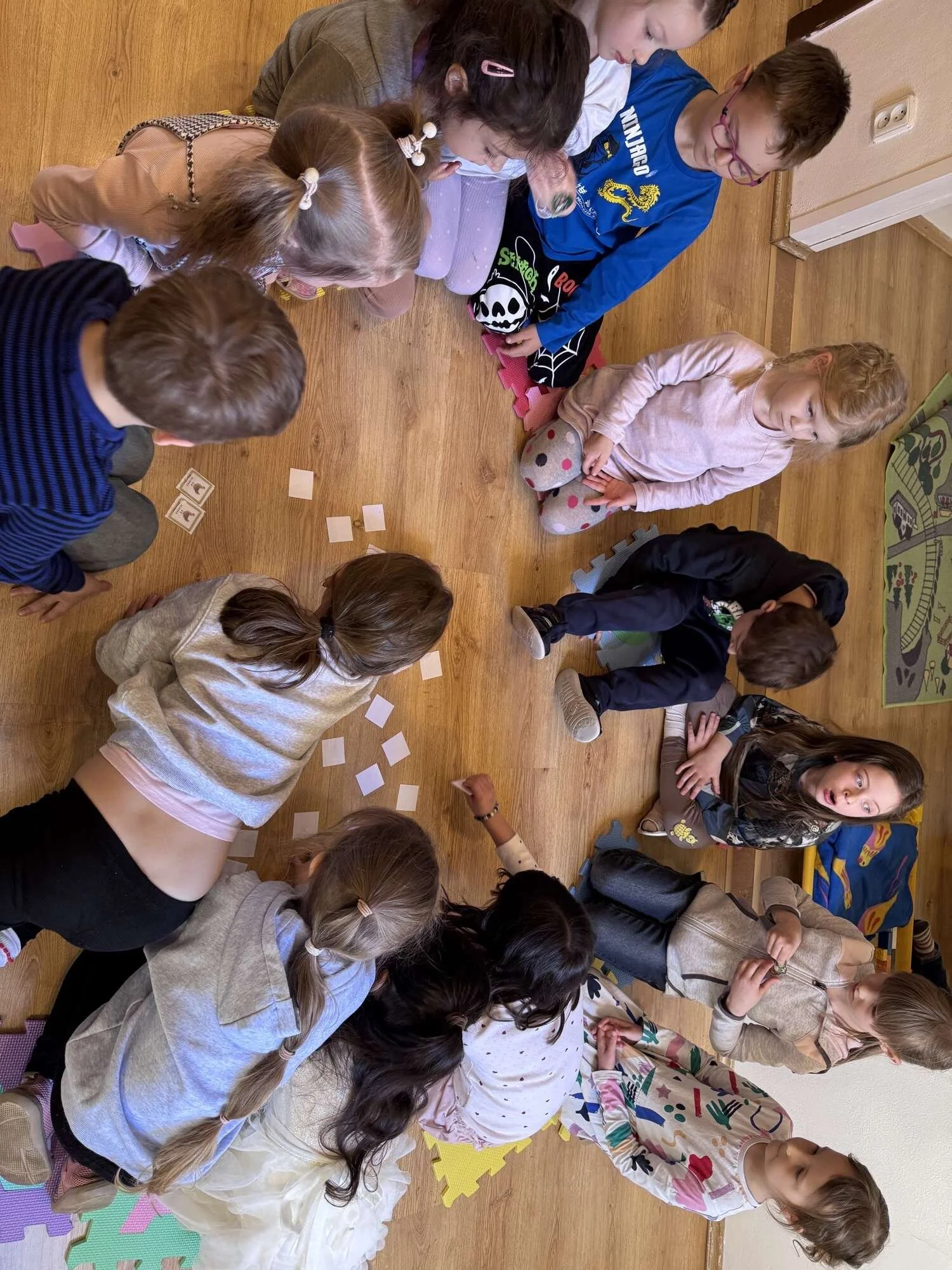 Children sitting and lying on a wooden floor, engaged in a group activity with small cards, some wearing colorful clothes and hair accessories.