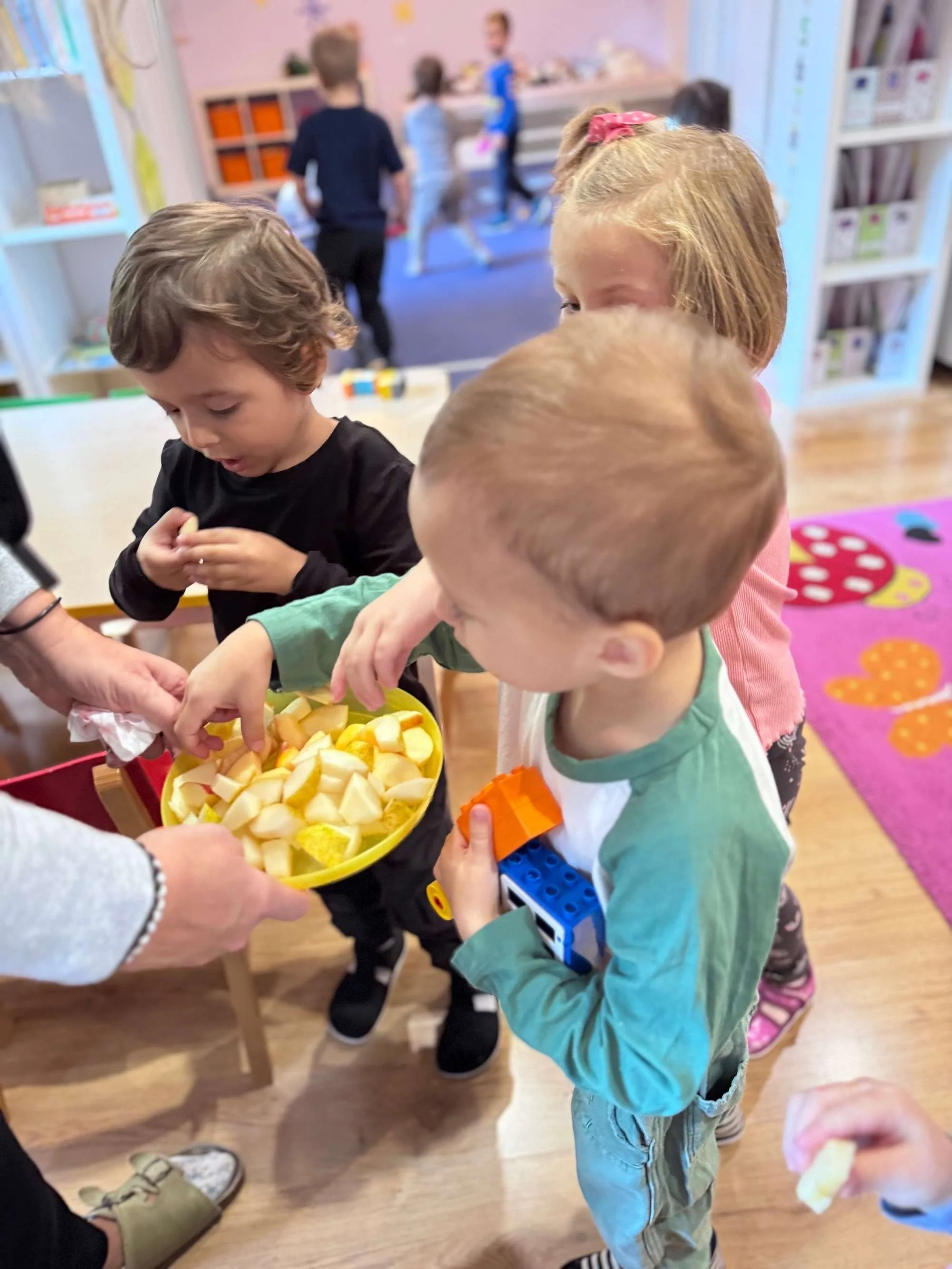 Children gather around a bowl of snacks, reaching for pieces, in a preschool or daycare setting.