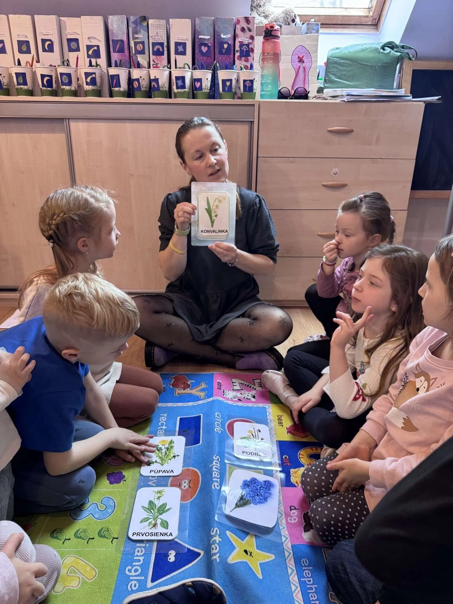 A teacher showing flashcards to a group of young children sitting on a colorful educational mat in a classroom.