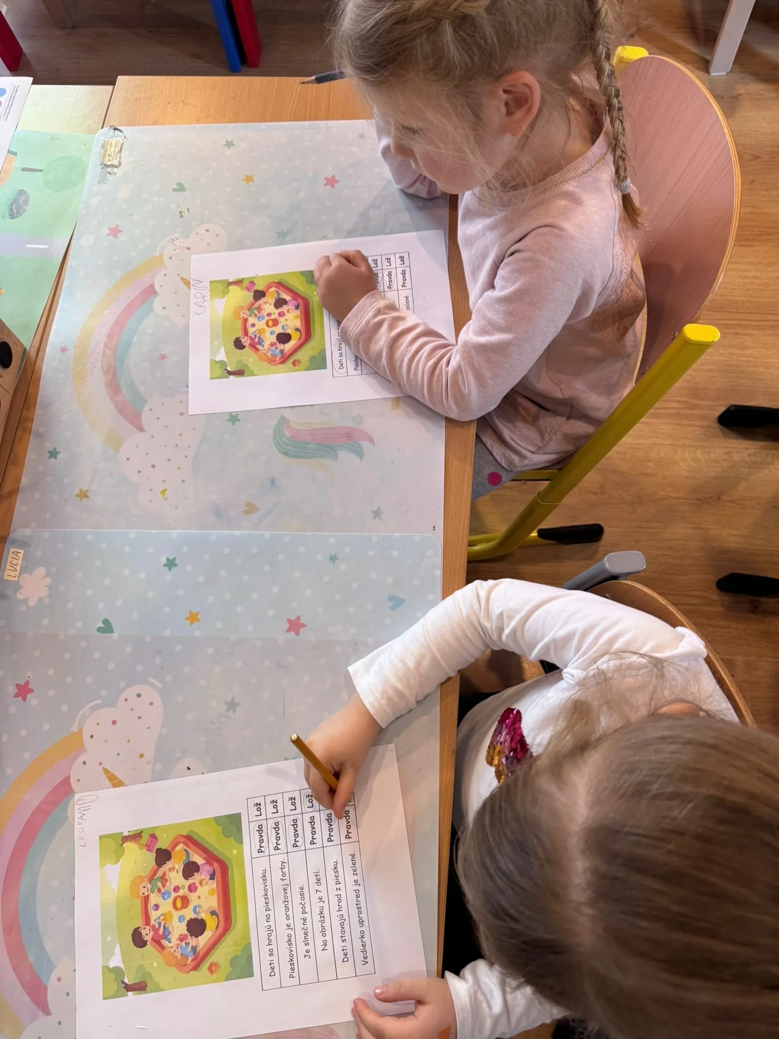 Two young girls sitting at a table, working on worksheets with a picture of a colorful playground and writing activities, in a classroom setting.