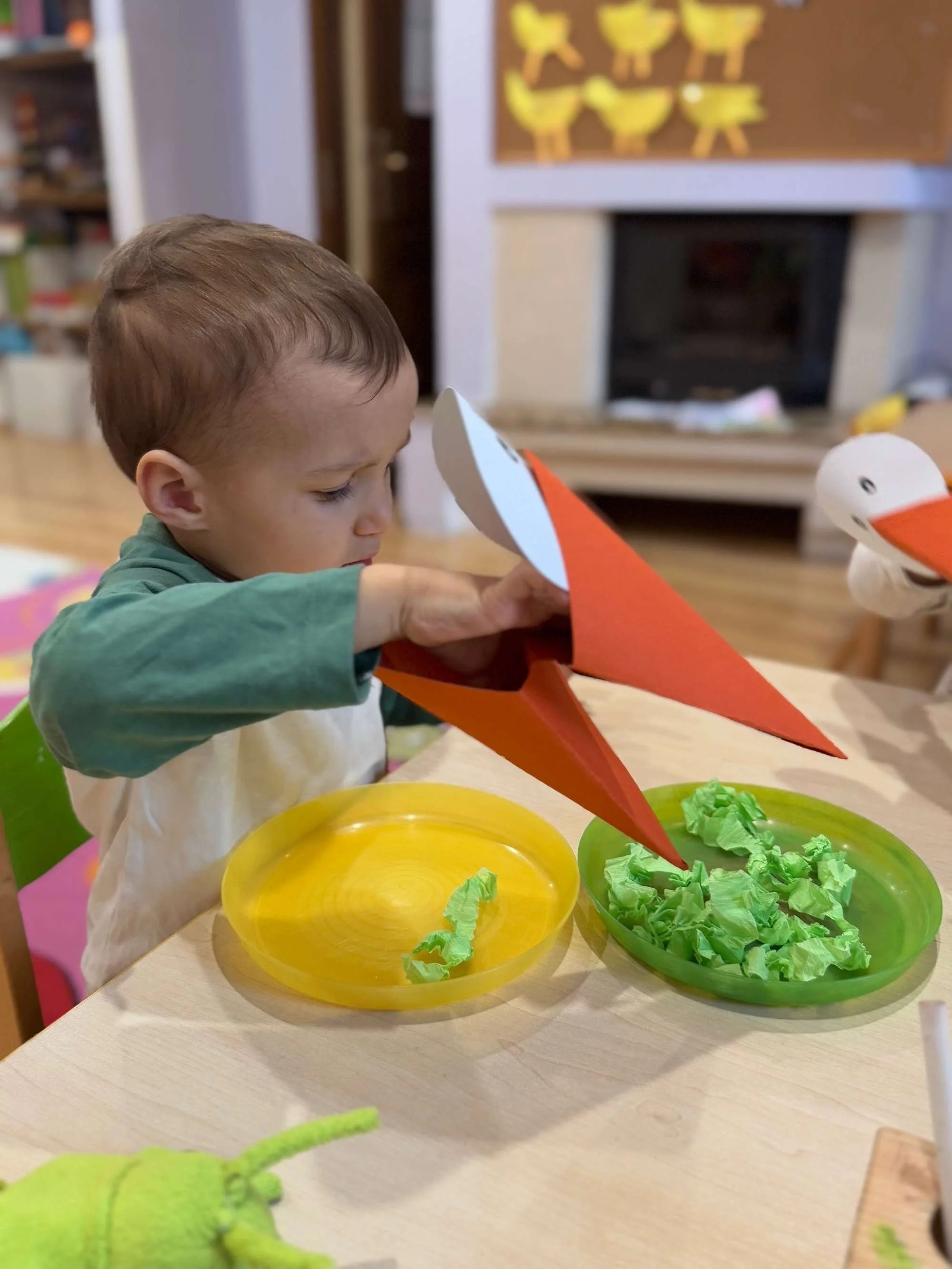 A young child playing with paper craft animals, including a stylized bird with a beak, at a table with green and yellow plates filled with crumpled green paper.