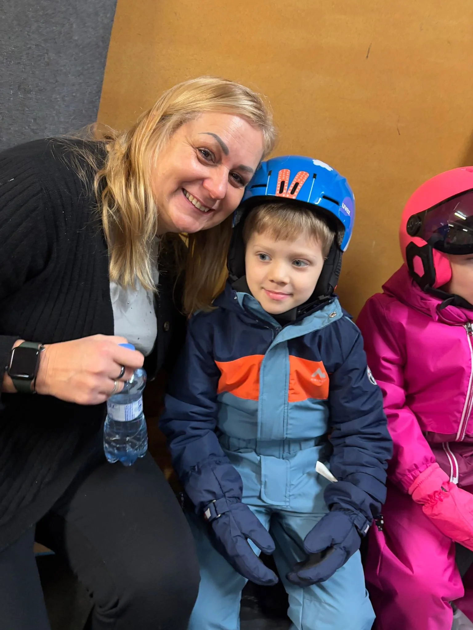 A woman smiling and holding a water bottle, sitting next to a young boy in a blue helmet and winter gear, with another child in pink winter gear and helmet partially visible on the right.