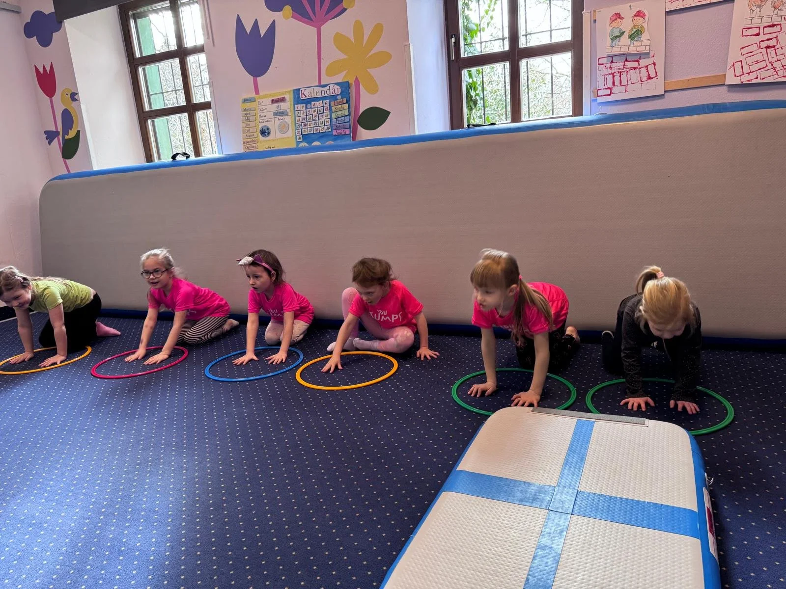 Six young girls crawling through hoops on a blue carpeted floor in a colorful classroom with windows and children's drawings on the walls.
