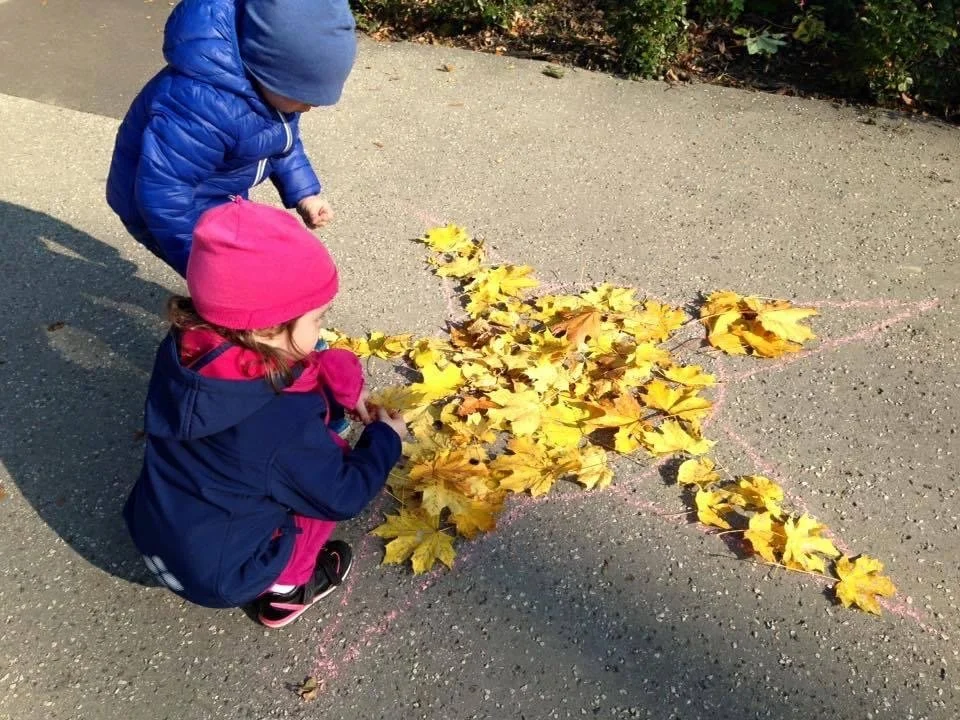 Two children in jackets and hats drawing with chalk on the sidewalk. They are creating a star shape outline filled with yellow and brown leaves.