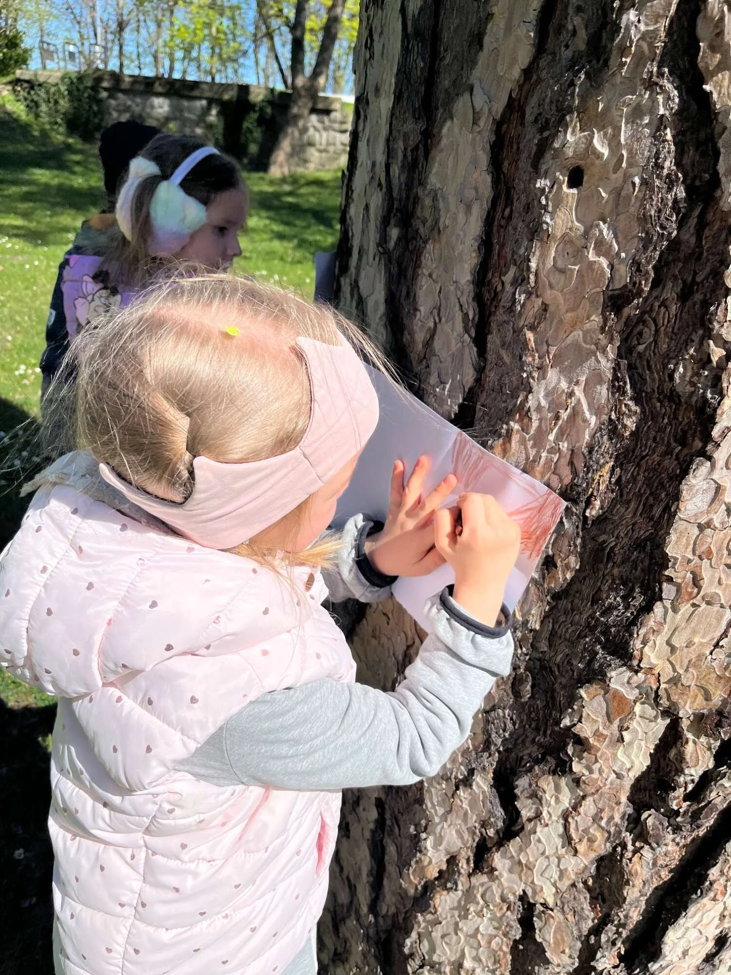 A young girl wearing a pink headband, gray sweatshirt, and a light pink vest is pinning paper to a tree trunk during an outdoor activity. Another girl in the background wears earmuffs and a floral jacket, observing in a park setting with grass, trees, and a stone wall.