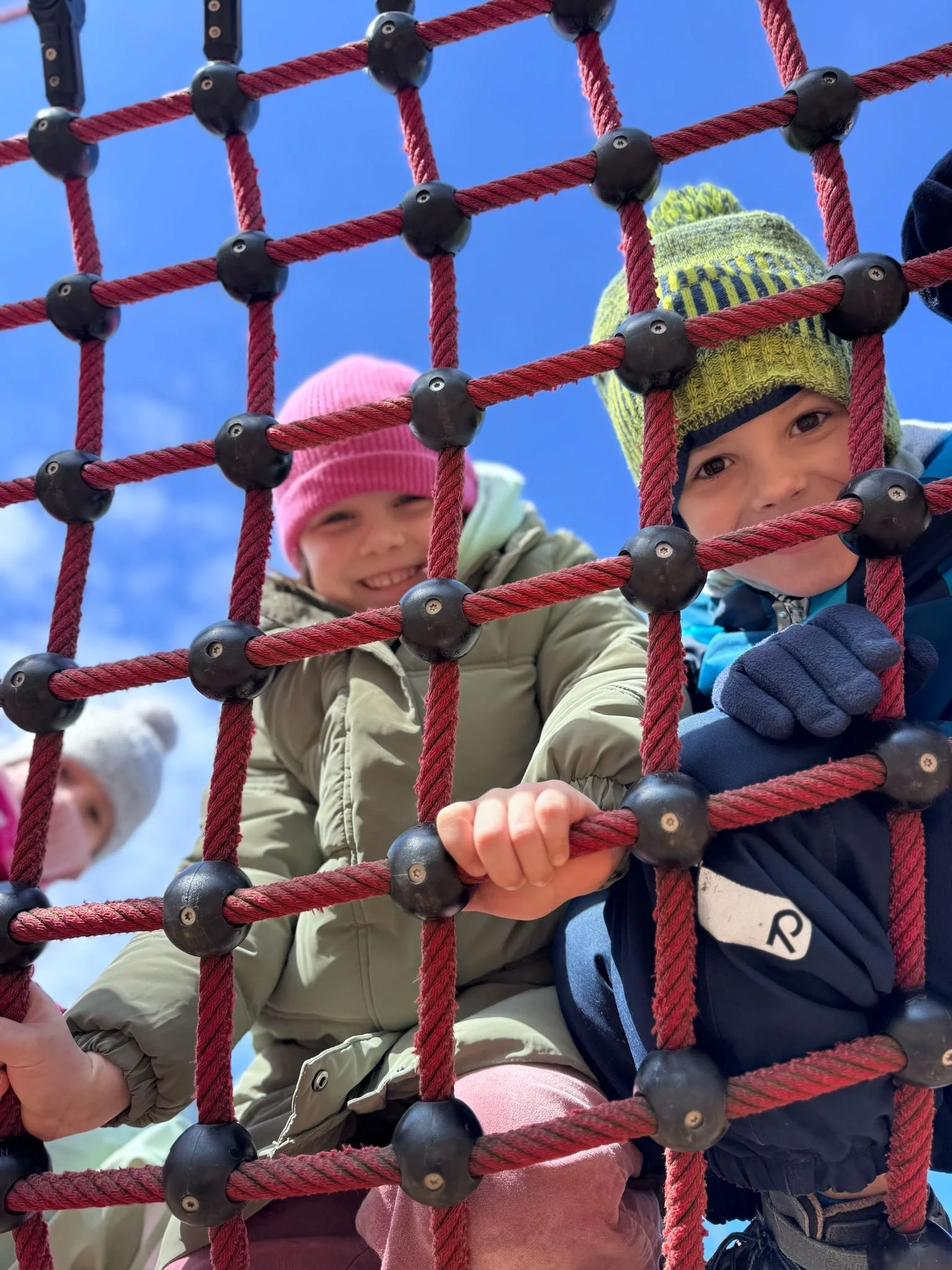 Two children climbing on a red rope net playground structure outdoors on a sunny day, with bright blue sky in the background.