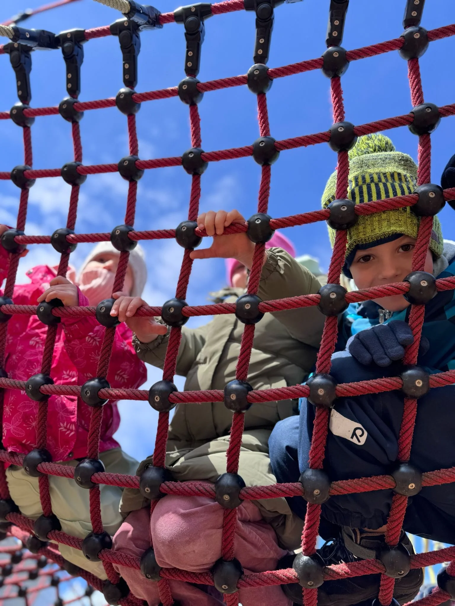 Children climbing on a red rope net structure outdoors, with blue sky and some clouds in the background.
