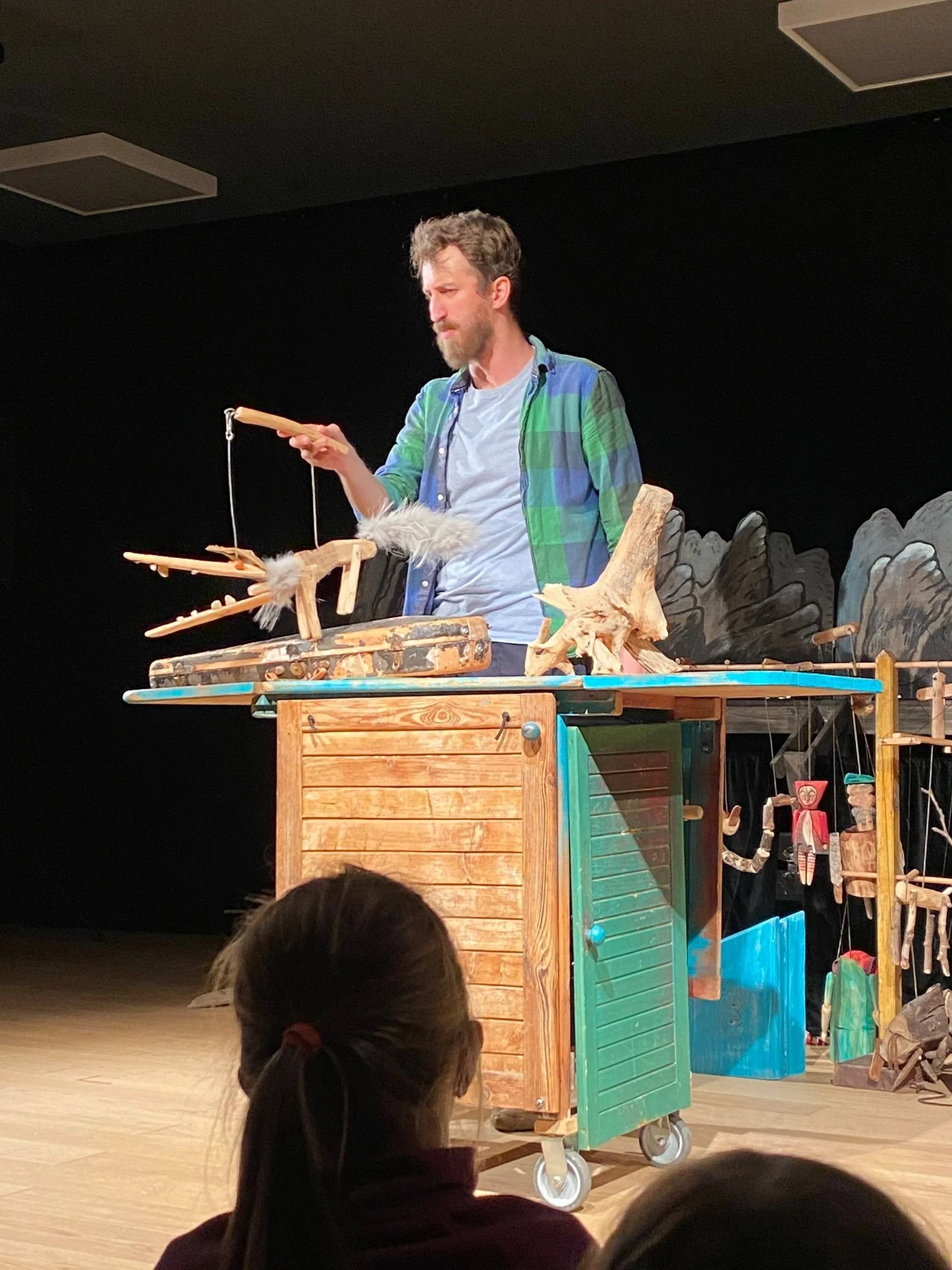 A man with a beard and curly hair performs a puppet show on stage, manipulating wooden puppets with furry details, with a backdrop of painted mountains and a wooden cart with colorful doors as part of the set.