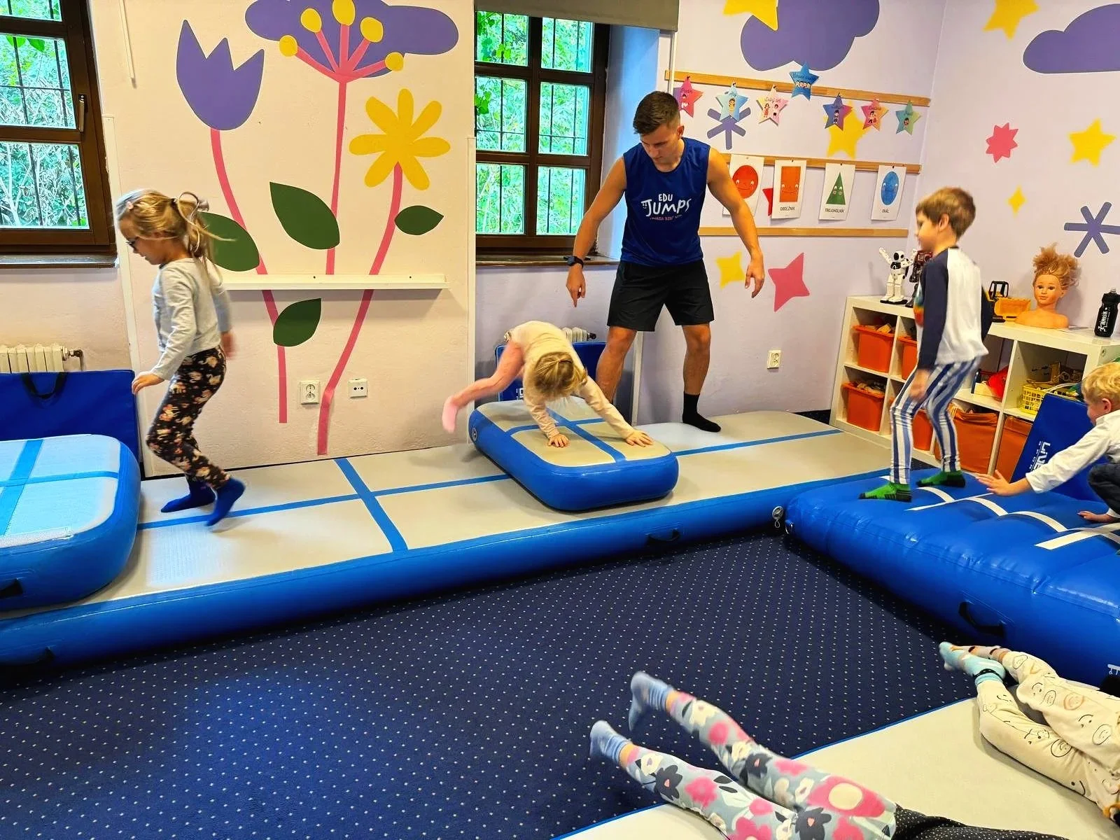 Children playing on an inflatable gymnastics area supervised by an adult in a colorful room with wall decorations of flowers and stars.