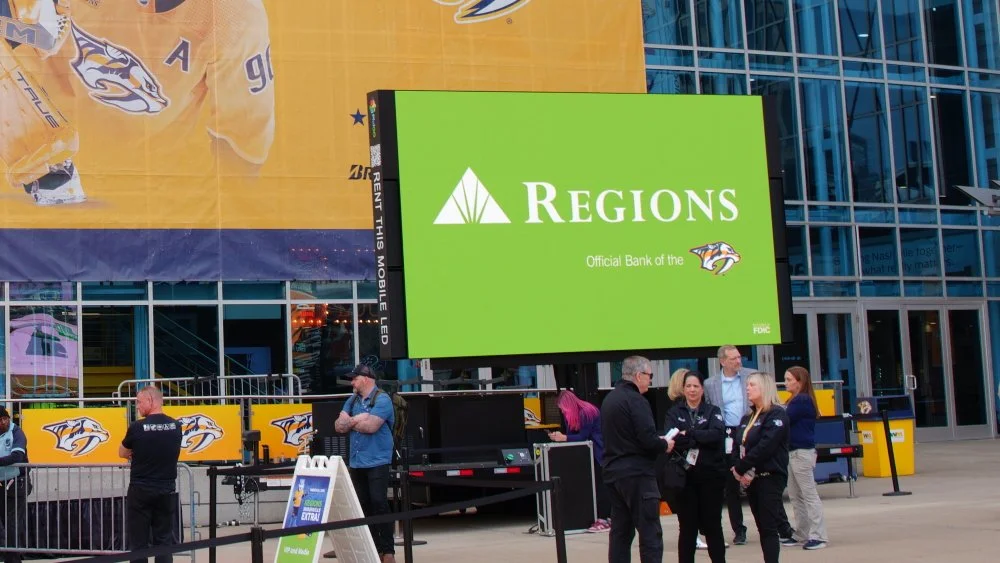 A large digital billboard outside a building displays an advertisement for Regions Bank with the Nashville Predators logo. People are standing and talking near the billboard, with some wearing Predators apparel and masks.