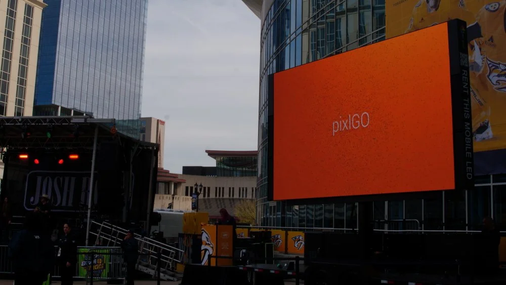Large orange digital billboard displaying "pixGO" in an urban outdoor setting with tall modern buildings and a small stage area.