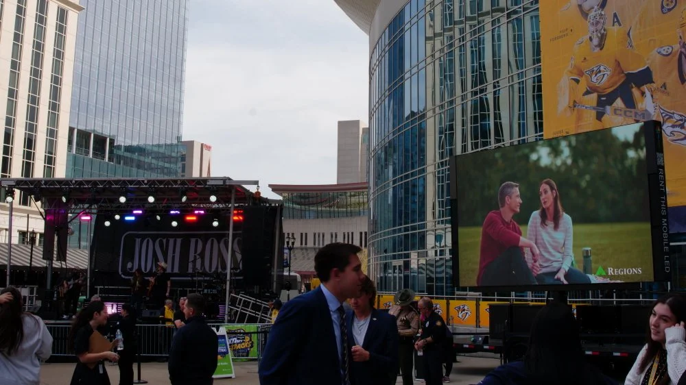 An outdoor event with a stage and large screen, surrounded by modern glass buildings, with people gathered and talking.