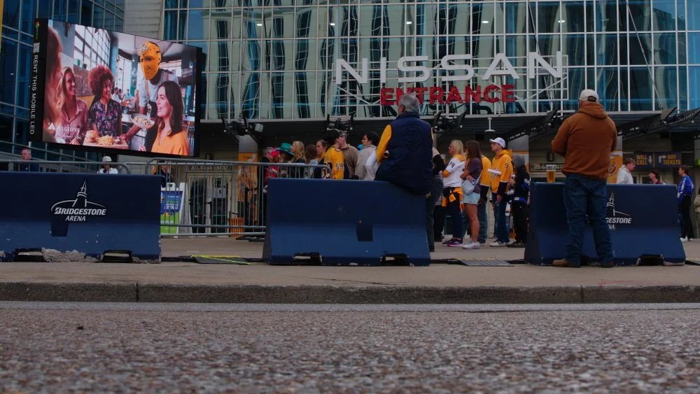 Crowd of people gathered outside the Nissan entrance at Bridgestone Arena, Nashville, Tennessee, with a large digital screen displaying images of people dining. Some are wearing team jerseys and casual clothing.