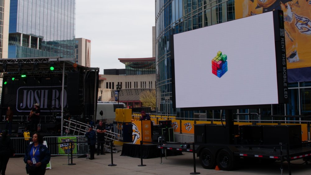 Outdoor stage setup with a large digital screen displaying a colorful 3D Rubik's Cube logo, in front of a modern glass building, with people, barriers, and a Tennessee Titans banner in the background.