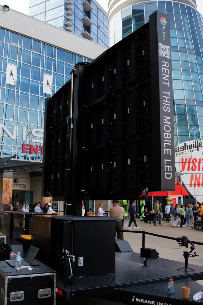 Large black mobile LED screen on a floating stage at a busy outdoor event in an urban area with modern glass buildings and crowds of people.