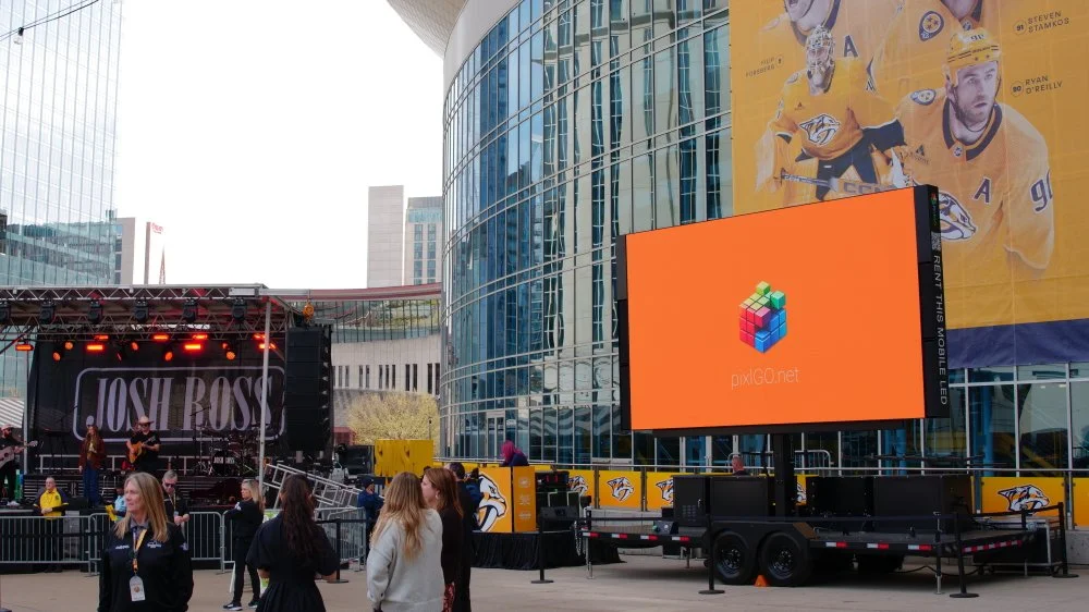 Outdoor concert with a stage, people, and a large digital screen displaying the pixelGO logo, in front of a modern glass building and a large hockey players poster.