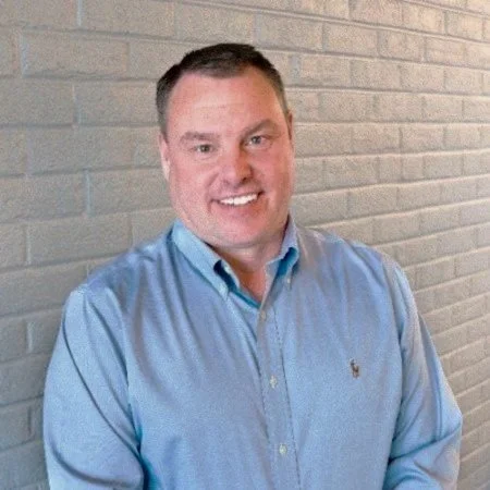 A man with short dark hair smiling and posing in front of a gray brick wall, wearing a light blue button-up shirt.