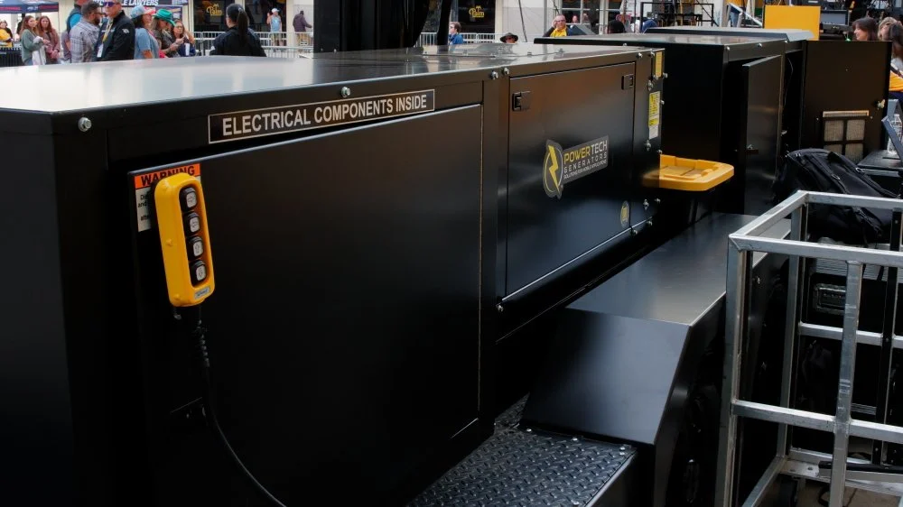 Large black generator with a warning label, a yellow remote control, and yellow accessories on top, set up outdoors at an event with people and tents in the background.