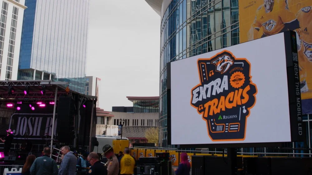An outdoor event with a stage and a large digital screen displaying a logo for 'Extra & Tracks' with information about a hockey team, Nashville Predators, in a city with modern glass buildings.