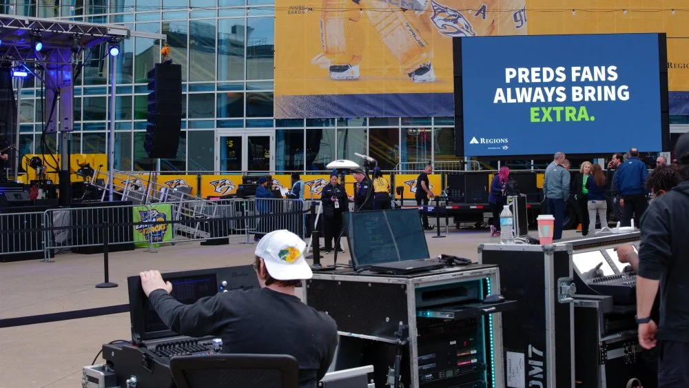 Outdoor event space with a large screen displaying 'PREDS FANS ALWAYS BRING EXTRA' and a crowd of people walking by. There is stage equipment and staff setting up in front of a modern glass building.