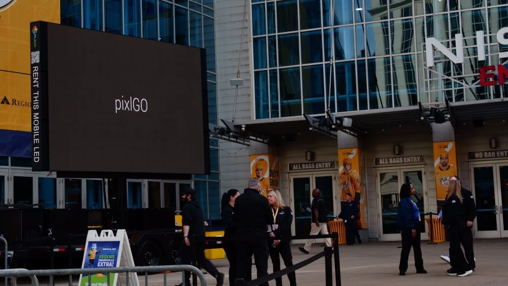 People standing outside an arena entrance with signs and large digital screen displaying the word 'pixiGO'.