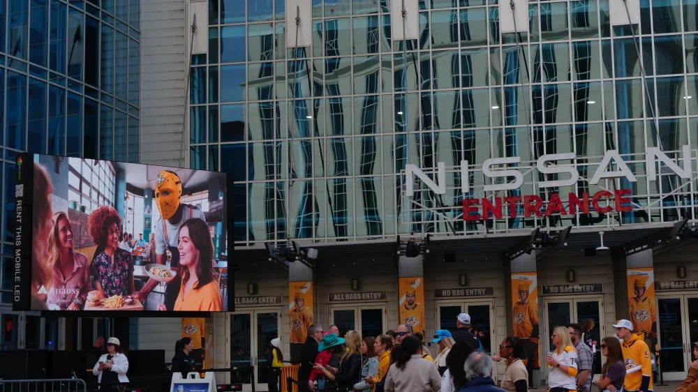 Crowd outside Nissan stadium in Nashville, with large digital screen showing people dining with a person wearing a hockey mask.