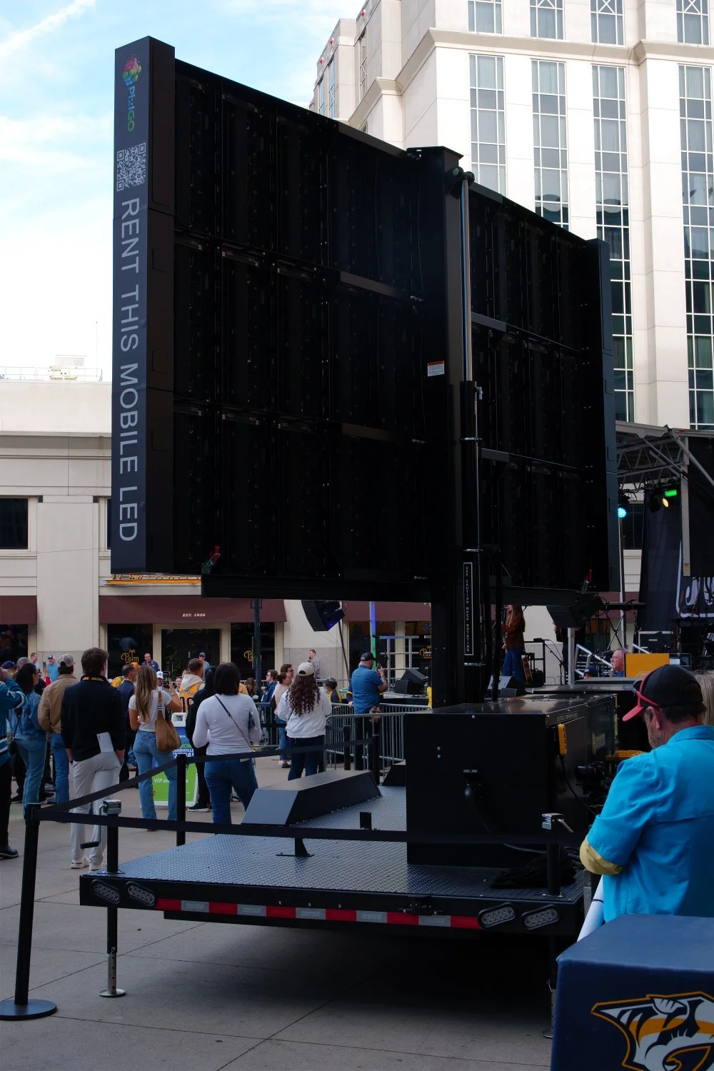 Large LED screen on a mobile display vehicle with people gathered around at an outdoor event, in front of tall buildings.