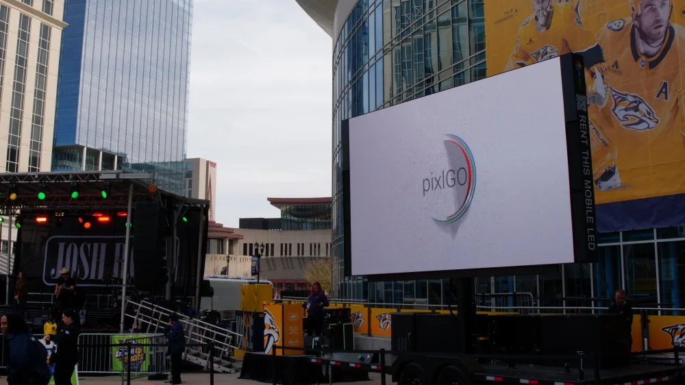 Outdoor stage setup with a large digital screen displaying the PiktoGO logo, with stage lights and people setting up, surrounded by banners and signs related to the Nashville Predators hockey team.