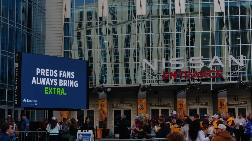 Crowd of fans outside Nissan Stadium, with a large digital signboard displaying a message about Preds fans bringing extra items. The stadium entrance features glass panels and signage for entrance and exit.