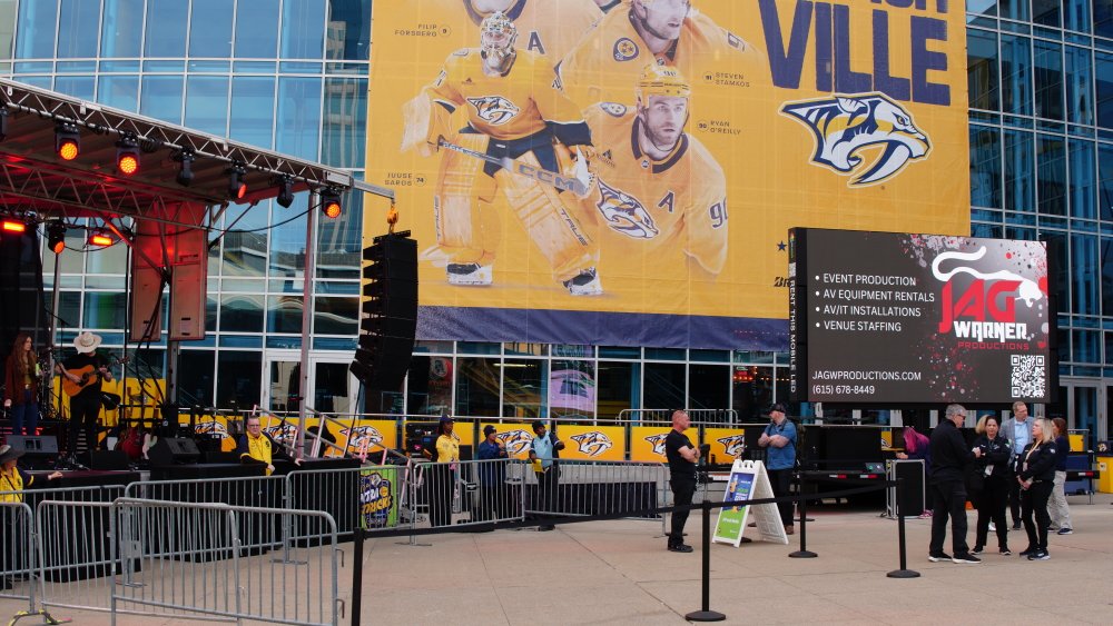Outdoor event setup in front of a glass building with a large yellow poster of Nashville Predators hockey players and team logo. A stage has musical instruments and equipment, with security barriers and a few people standing around.