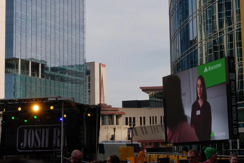 Outdoor concert in an urban setting with stage lights, a large digital screen displaying a woman, tall glass buildings, and people in the crowd.