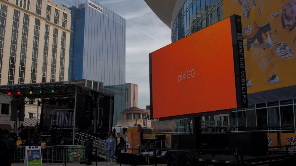 An outdoor scene in a city with tall buildings featuring glass facades. A large red digital screen displays the word 'pixiGO.' A stage with a black canopy and lights is set up nearby, with people walking around.