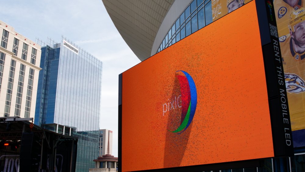 A large digital billboard displaying an orange background with a partially visible 'pixel' logo. The billboard is situated in an urban area surrounded by tall buildings, including a Bridgestone building, and features a vertical strip of text on the r