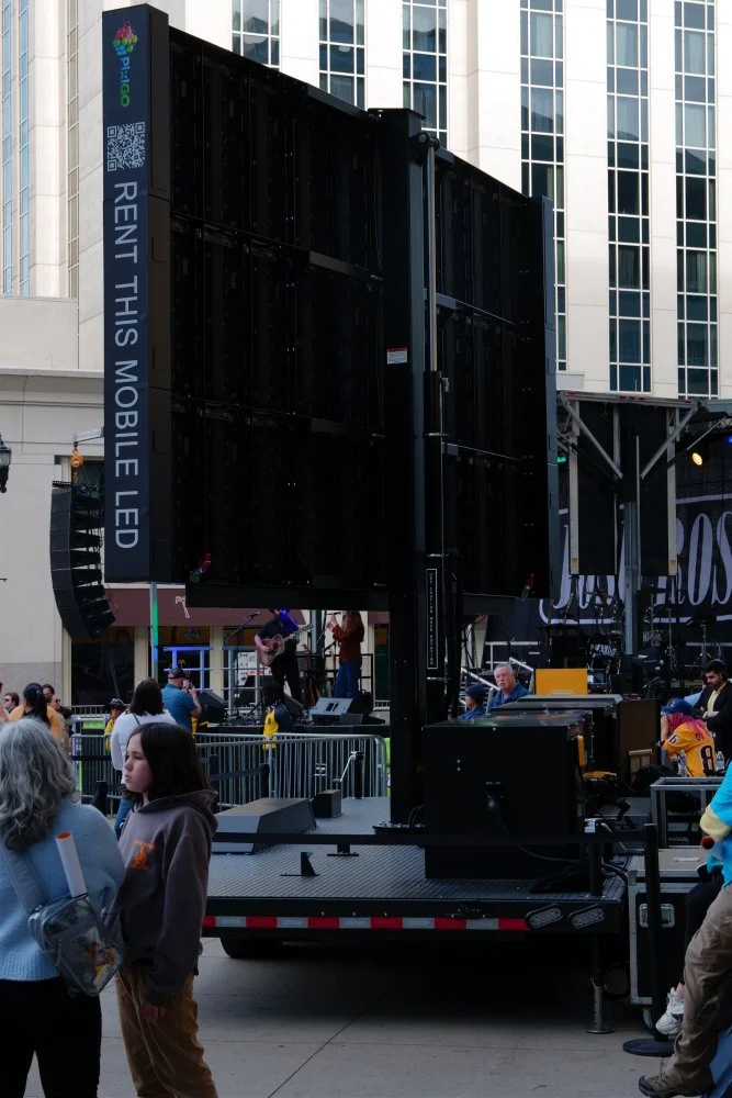 Large black mobile LED screen on a truck at an outdoor event with people gathered around, some watching a performance on stage in the background, in front of tall glass buildings.