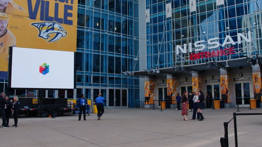 Exterior of Nissan Arena with large glass windows, banners, and a digital billboard, people walking outside.