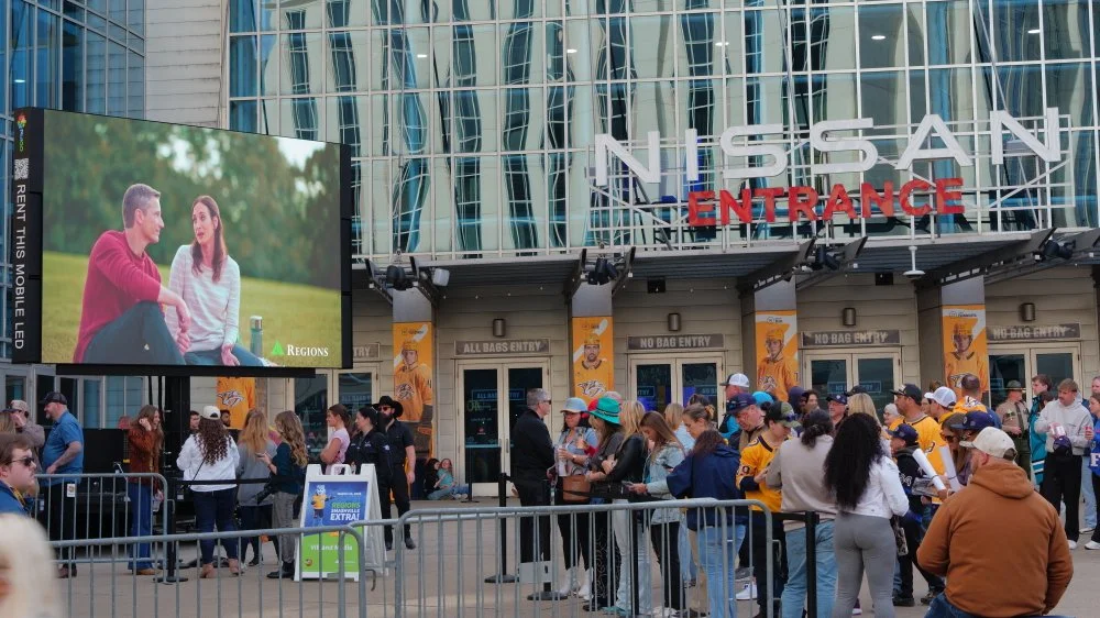 Crowds lining up outside Nissan Arena in front of a large LED screen showing two people sitting outdoors, with a backdrop of trees. The arena has a modern glass facade and prominent Nissan Entrance sign. There are signs indicating 'No Bag Entry' and 