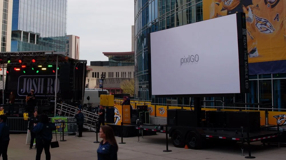 Outdoor event stage with green and red lights, banners, and a large LED screen displaying "pixiGO" in a city square surrounded by modern buildings and security personnel.