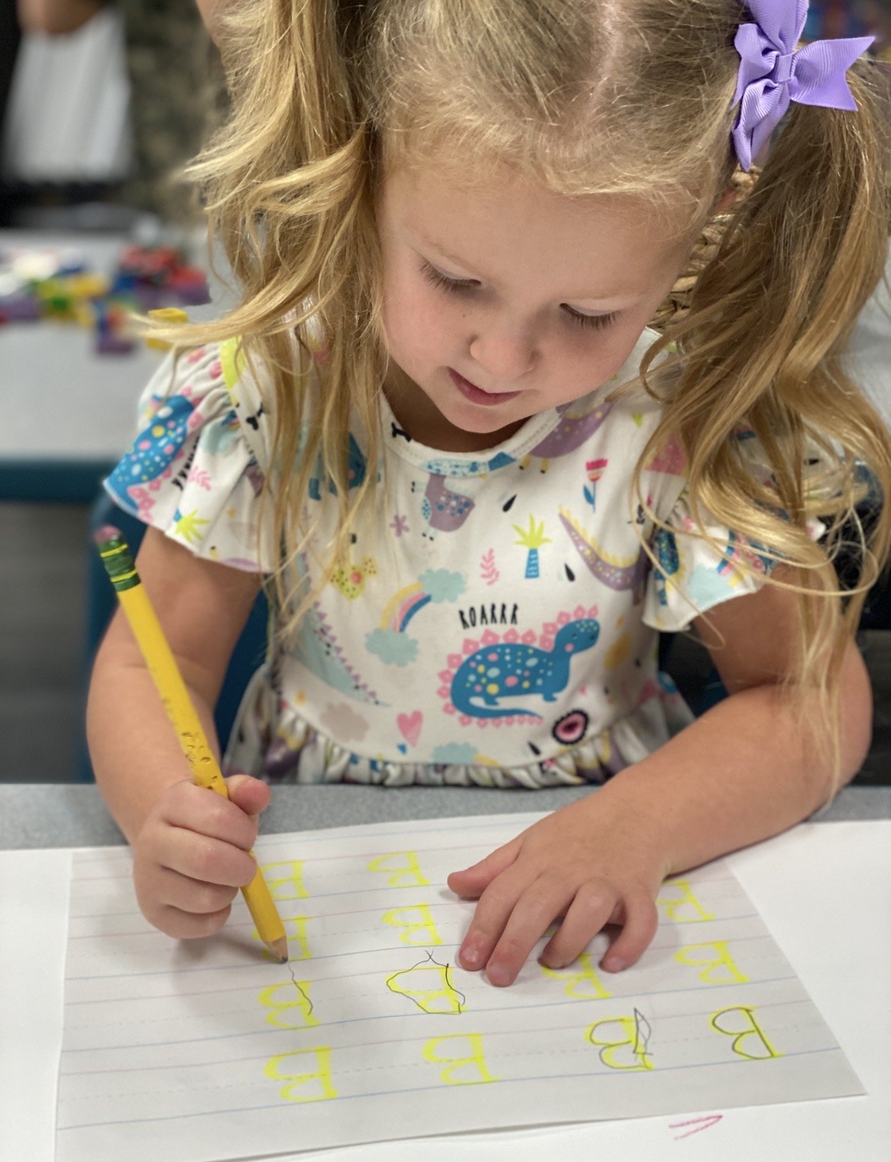 A young girl with curly blonde hair, wearing a colorful dress with dinosaurs and rainbows, is sitting at a table writing the letter 'B' repeatedly in yellow on a lined sheet of paper with a yellow marker.
