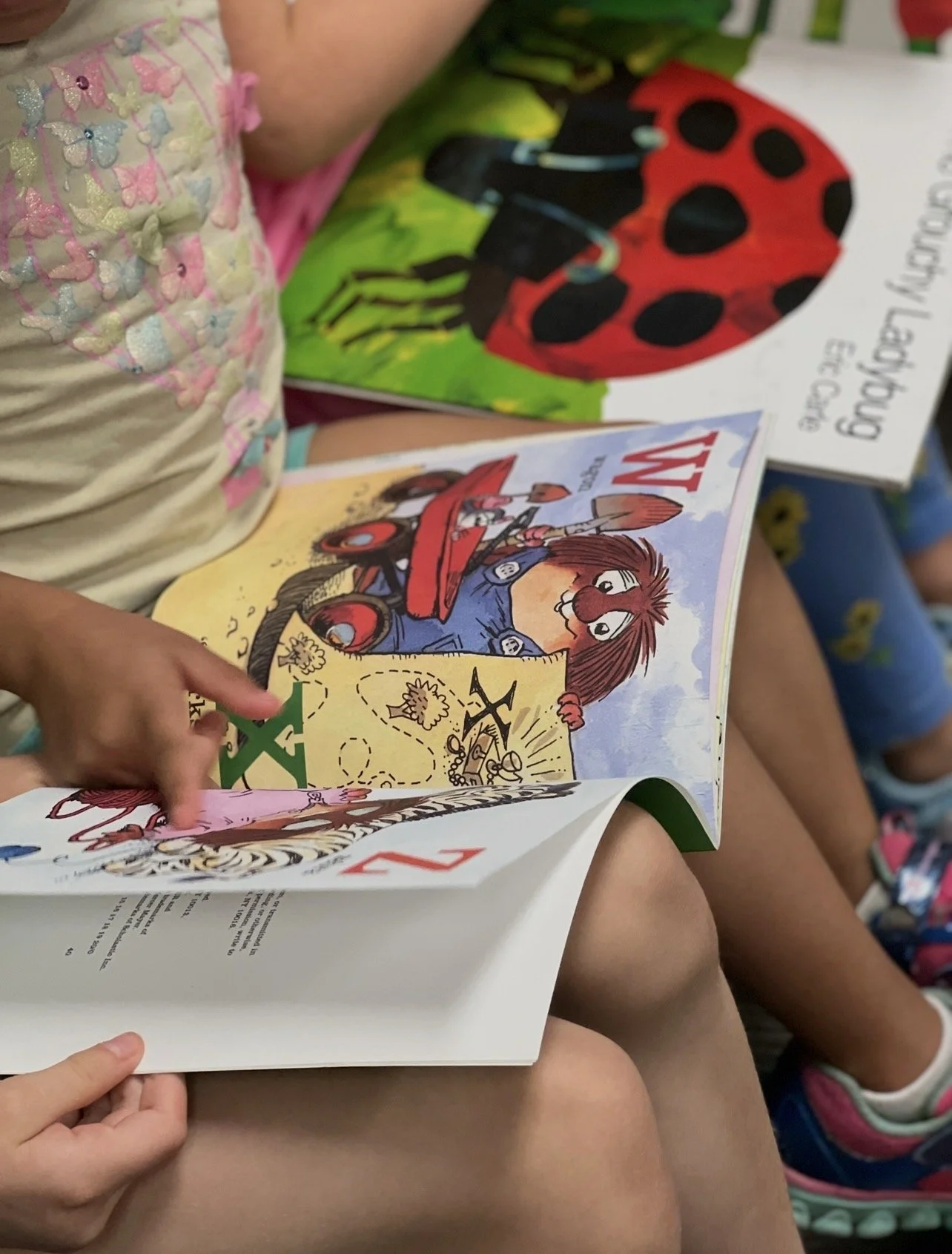 Children sitting and reading colorful picture books, one with a Robot and animal story, and the other with a ladybug and nature theme.