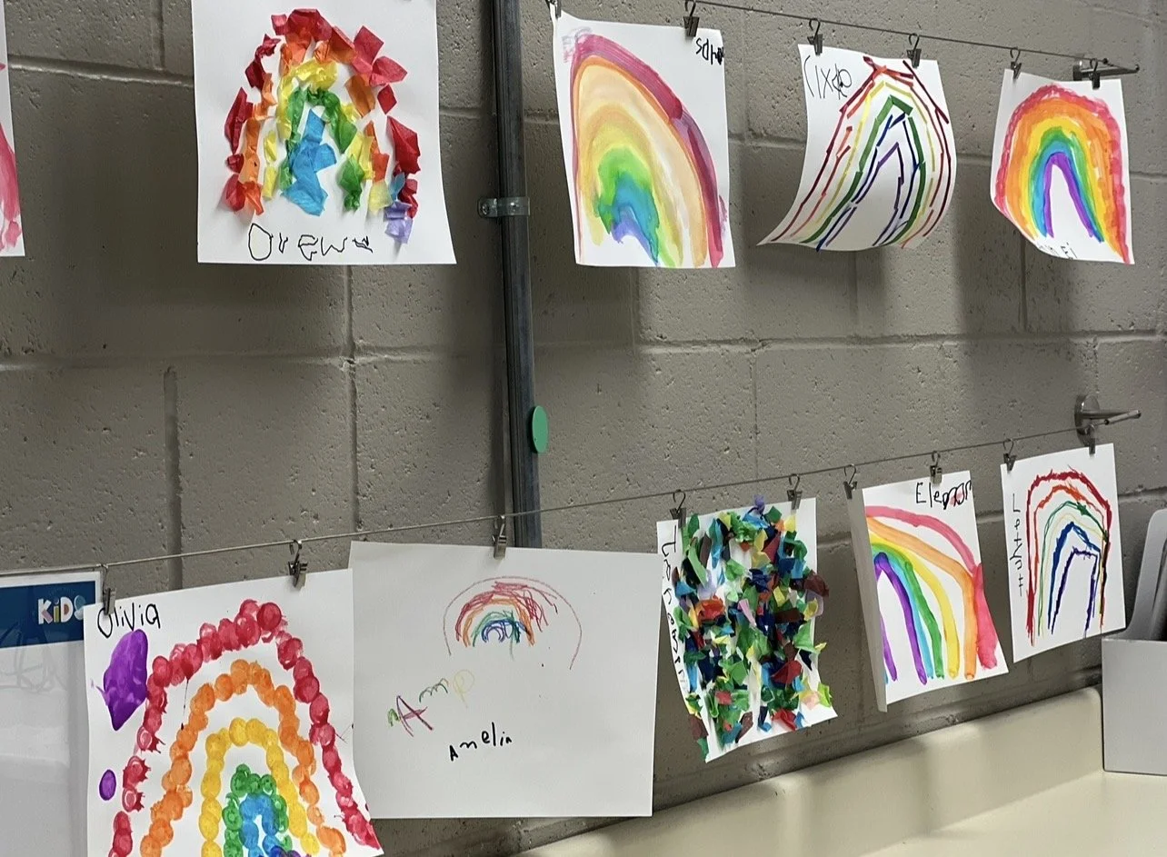 Children's colorful rainbow artwork hanging on a wire with clips, created with various art supplies including torn tissue paper, markers, and paint, labeled with children's names.
