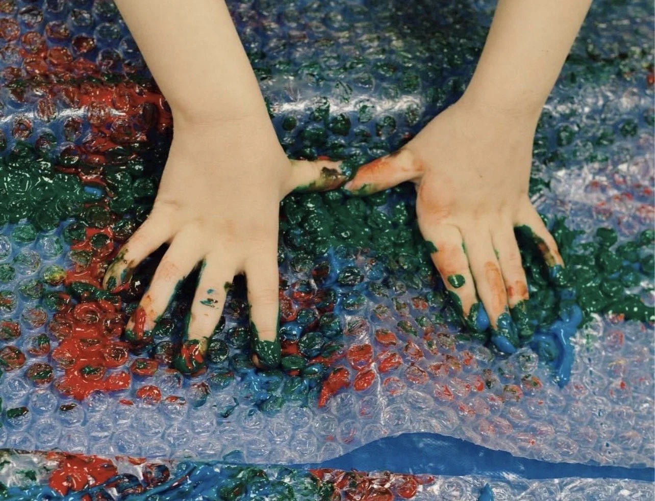 Hands covered in green, blue, and red paint pressing down on a bubble wrap surface with colorful paint blobs at Adventure Academy child care in Burbank.