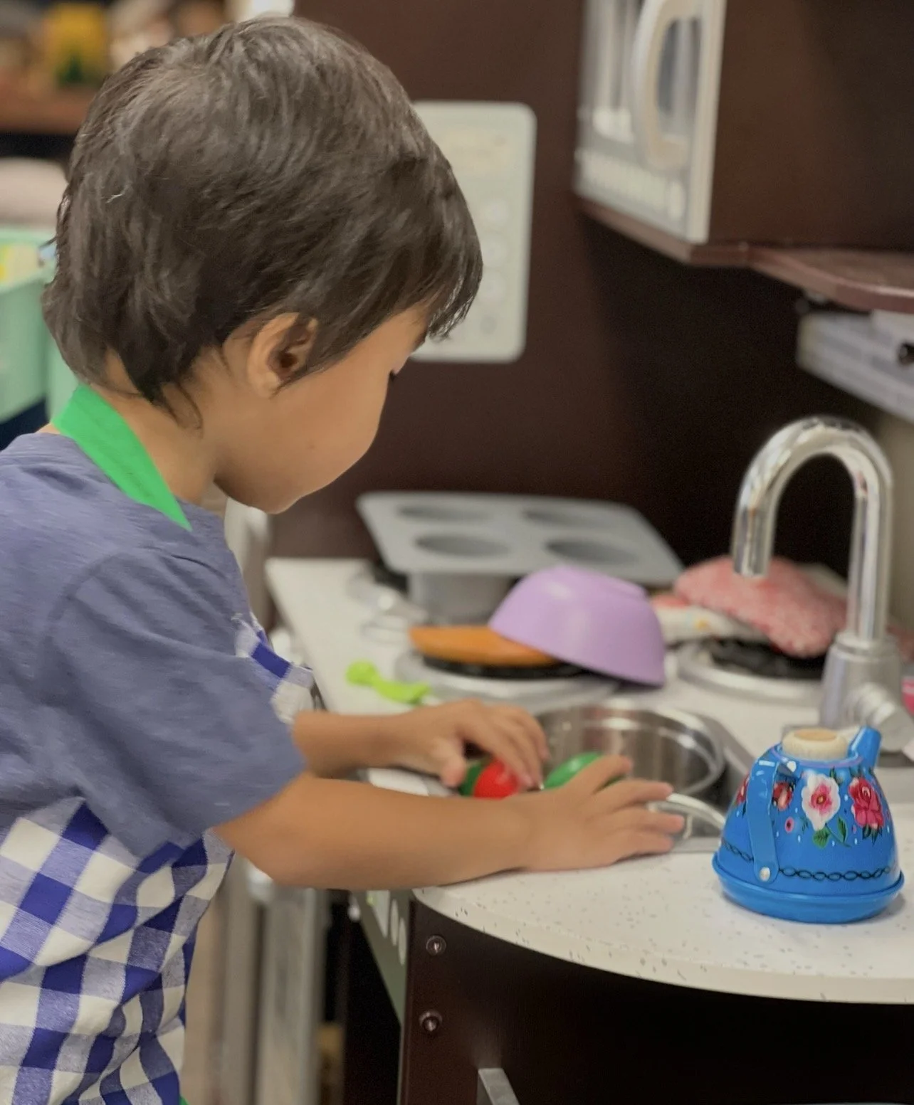A young boy wearing a blue apron with white checkered pattern, standing at a kitchen counter, washing plastic toy vegetables in a sink.