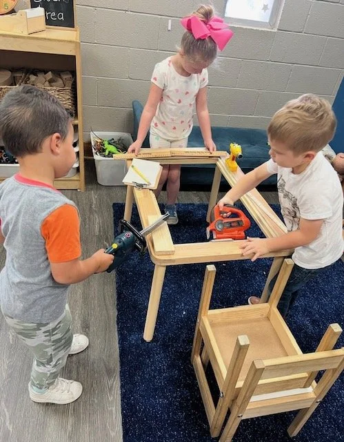 Three children play with wooden blocks and toy tools in a classroom, building a structure.