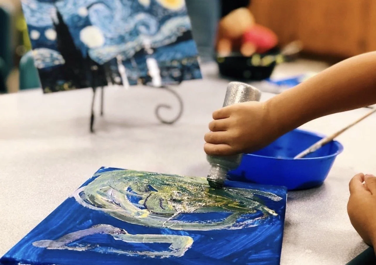 Child's hand using a silver squeeze bottle to paint an abstract design with green, white, and yellow paint on a blue canvas. Art supplies and a bowl of paint are visible on the table.