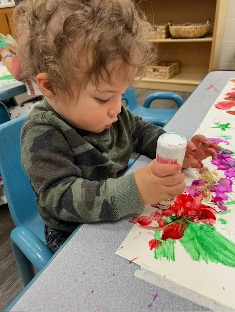 Young child with curly hair in camouflage sweater painting with colorful markers on paper at a table in a classroom.