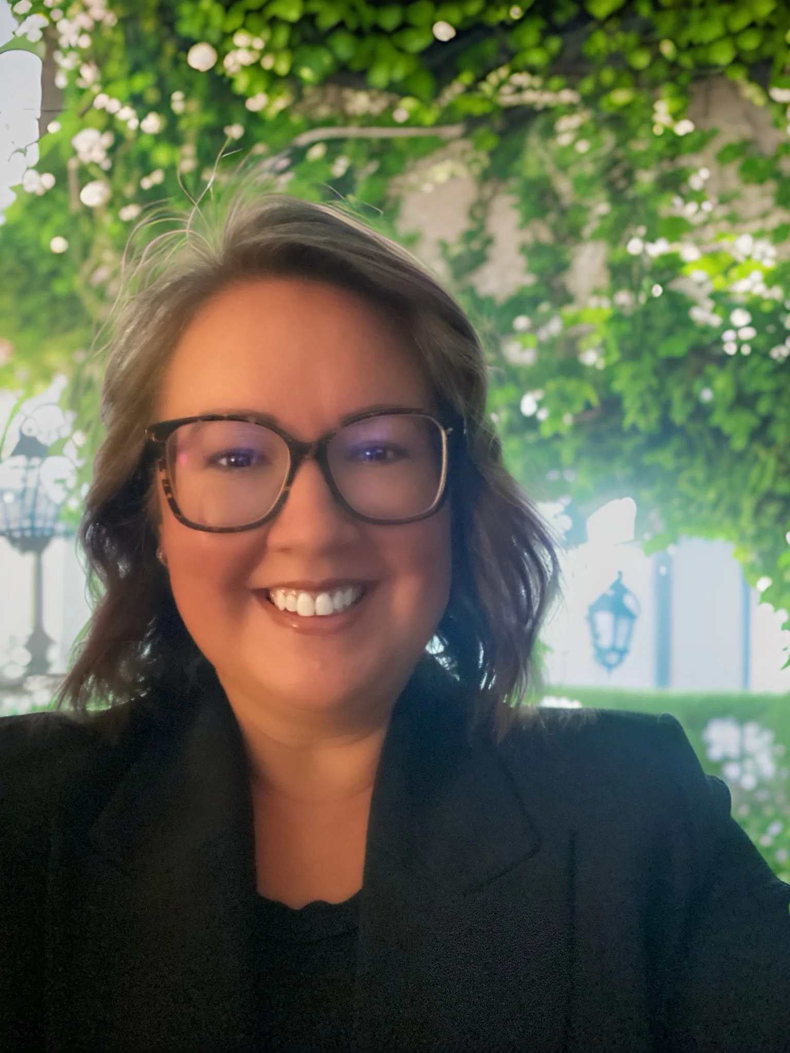 A woman with shoulder-length hair, glasses, and a black blazer smiling outdoors with green foliage and decorative lamp posts in the background.